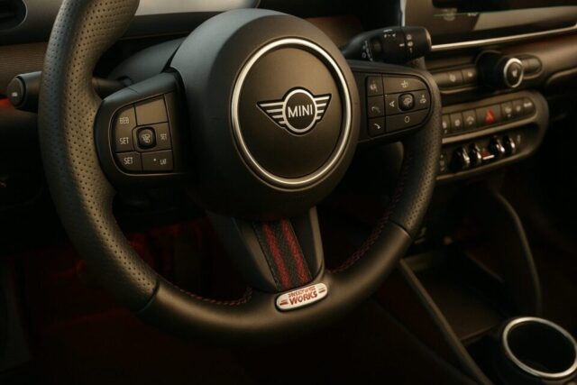 レザー巻きステアリングホイールが特徴のMINI John Cooper Worksのインテリア。赤いステッチが施されたスポーティなデザインで、ホイール中央にはクラシックなMINIロゴ、下部には「John Cooper Works」のメタルエンブレムが光る。ドライビングモードやクルーズコントロールなどの操作ボタンが集約され、機能性とスタイルが融合したデザイン。