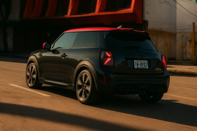 黒いボディに赤いルーフが特徴のMINI John Cooper Worksハッチバックが都市の街並みを背景に走行中。ユニオンジャックモチーフのテールライトやレッドキャリパー付きのスポーツホイールが際立ち、スポーティでアグレッシブなデザインが強調されている。