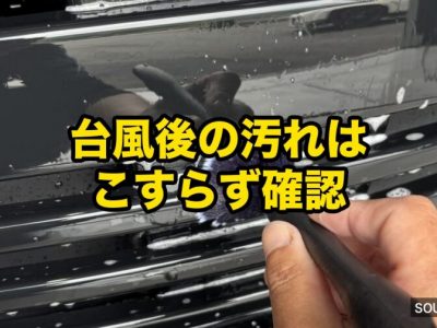 Person wipes a car's front grille with a cloth; yellow Japanese tip overlay says '台風後の汚れはこすらず確認' ('After a typhoon, check dirt without rubbing').